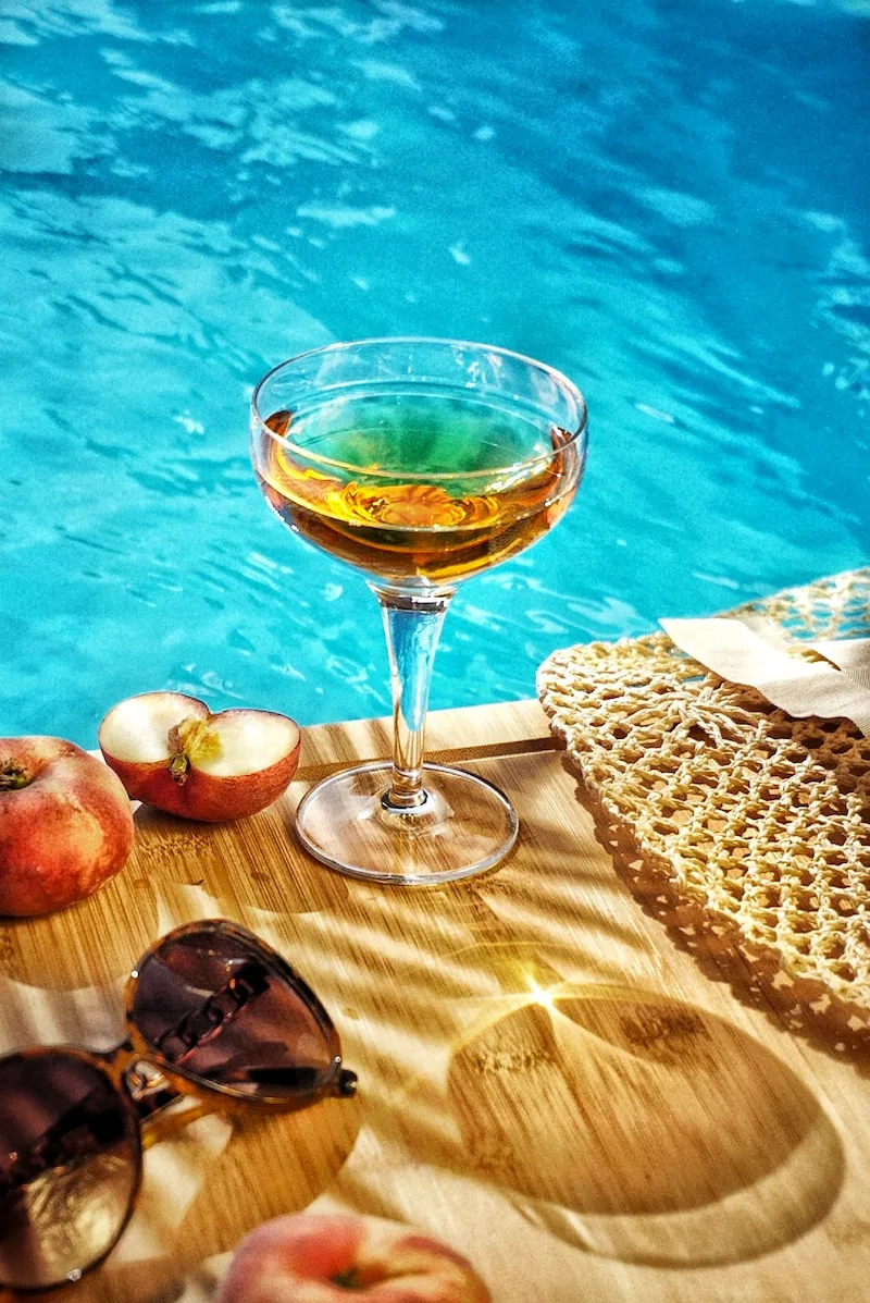 Erfrischendes Sommergetränk im Glas am Pool mit Pfirsichen und Sonnenlicht, fotografiert von Studio Visionelle.