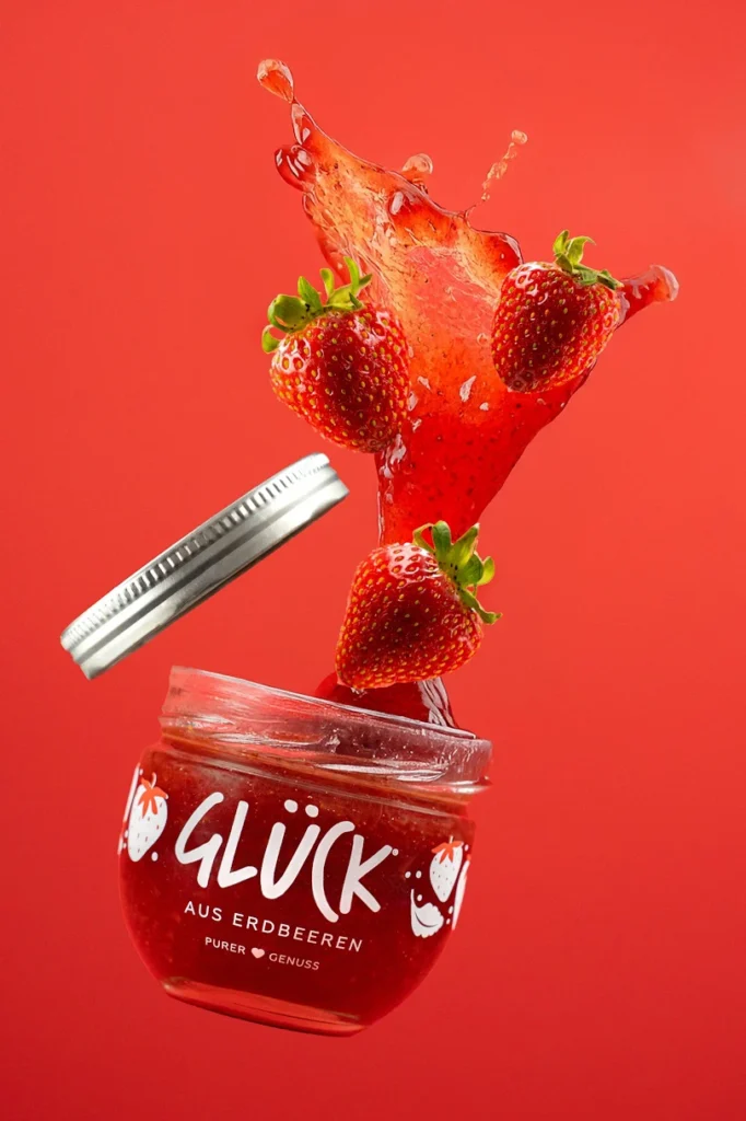 Fliegende Erdbeermarmelade vor rotem Hintergrund mit spritzenden Erdbeeren, dynamisch fotografiert von Studio Visionelle.