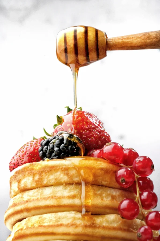 Close-up von Pancake-Stapel mit frischen Beeren, übergossen mit Honig von einem Löffel, fotografiert von Studio Visionelle.